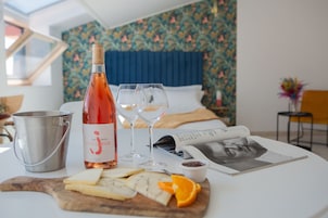 Un desayuno ordenado con una botella de Cuvée Bois, dos vasos, un cubo metálico, queso, una naranja y una revista sobre una cama.