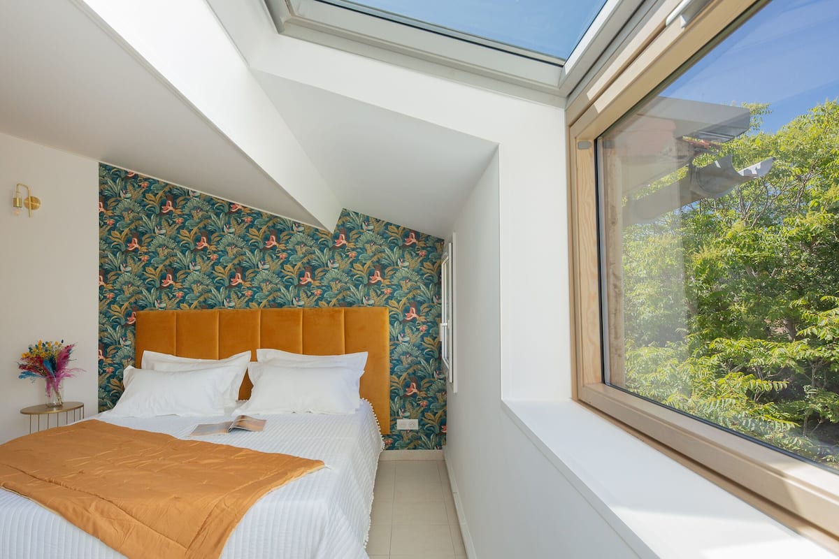 A bedroom with a skylight, a bed with white bedding, a floral wallpaper, and a view of trees through a window.