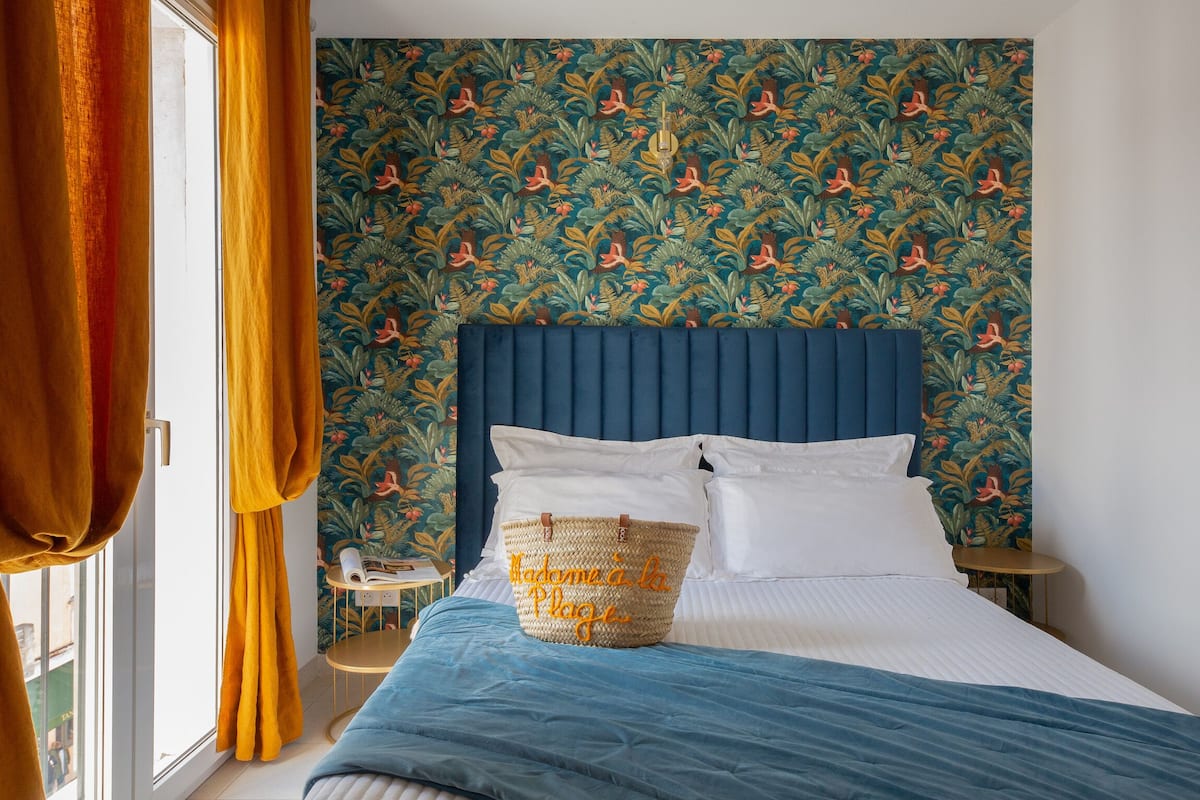 A bedroom with a blue headboard, a bed with white linens, a blue blanket, and a woven basket with text on it.
