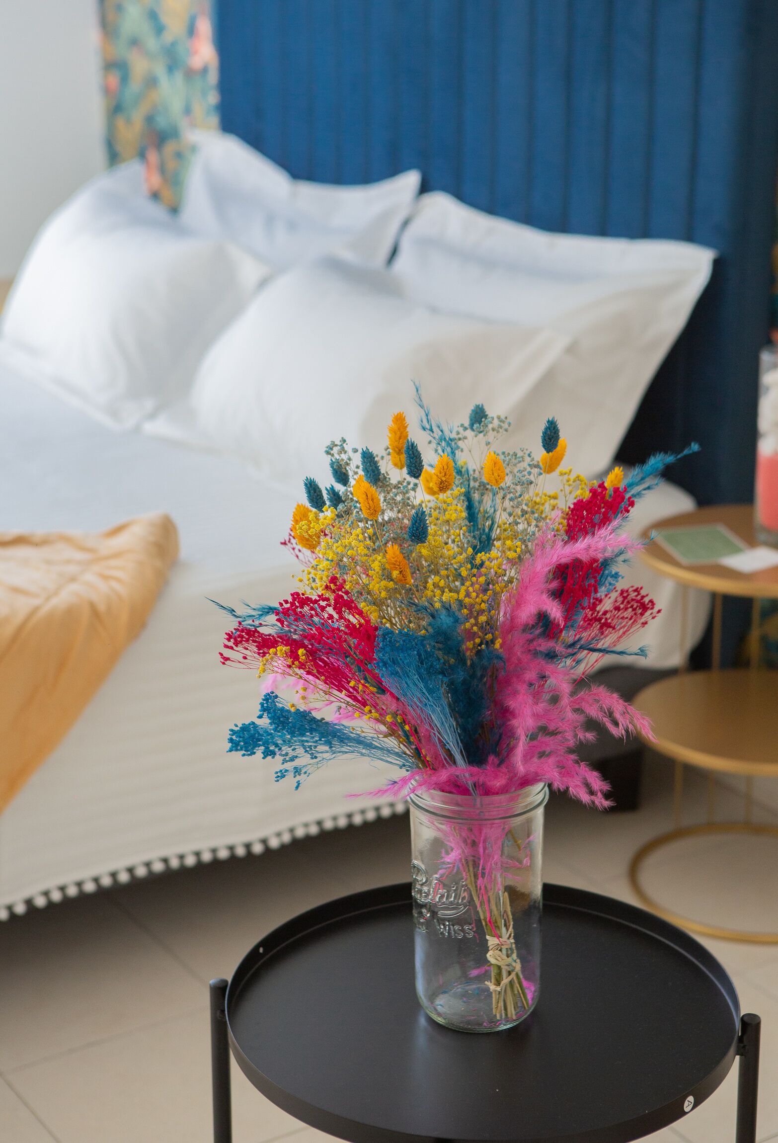 Una cama con sábanas blancas y un florero con flores coloridas sobre una mesita redonda.