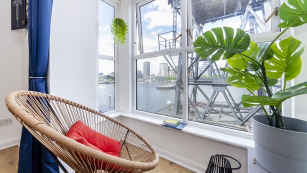 A room with a wicker chair, a red pillow, a potted plant, and a view of a bridge and water. - Homely 2-bed Apartment in London (London)