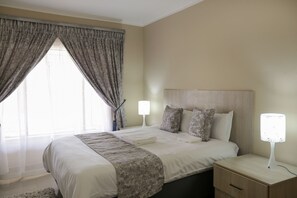 Una cama bien hecha con cabecera, una mesita de noche con lámpara y una ventana con cortinas.