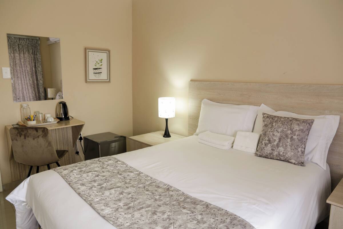 Ein Hotelzimmer mit einem großen Bett, einem Nachttisch mit Lampe, einem Schreibtisch mit Spiegel und einem gerahmten Bild an der Wand.
