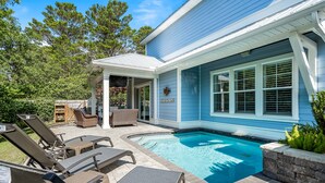 Uma área de piscina com espreguiçadeiras, um alpendre coberto e uma casa com revestimento azul e detalhes brancos.