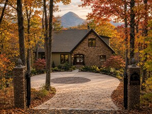 Une maison en pierre avec une allée circulaire, entourée du feuillage automnal et avec une montagne en arrière-plan.