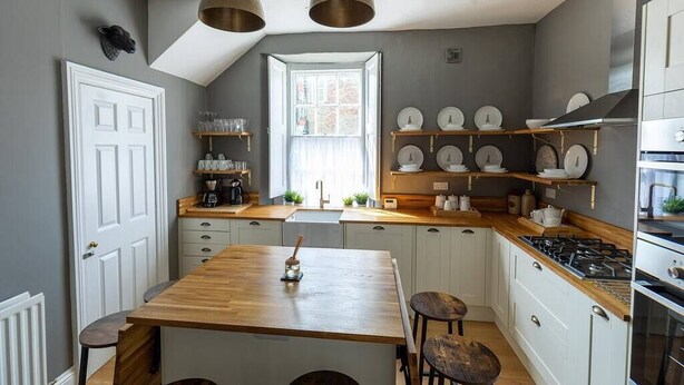 A kitchen with a wooden island, white cabinets, and a gas stove. There are two pendant lights, a window, and shelves with dishes and kitchenware. - DUNDAS COTTAGE - 3 Bed in Richmond North Yorkshire (Richmond)