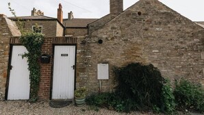 A stone building with two white doors, a brick wall, and a small garden area with plants and a potted plant. - DUNDAS COTTAGE - 3 Bed in Richmond North Yorkshire (Richmond)