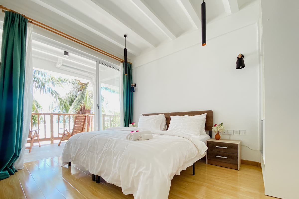 Un dormitorio espacioso con una cama grande, suelo de madera y un balcón con vistas a palmeras.