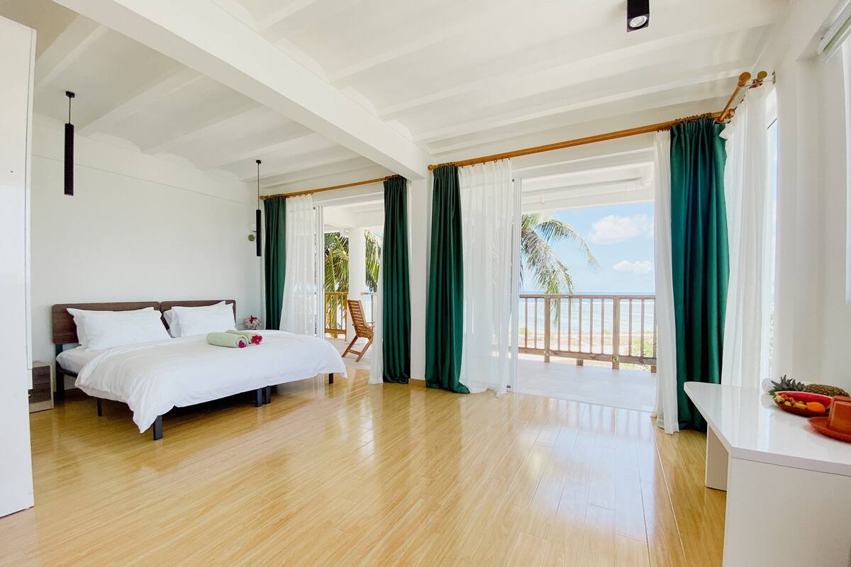 Una habitación de hotel con cama, suelo de madera, balcón con cortinas y vista a la playa.