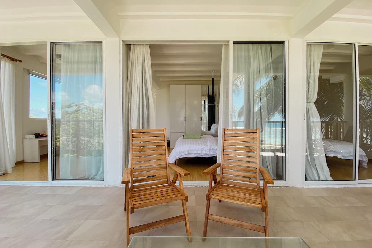 Una habitación con una mesa de vidrio, dos sillas de madera, una cama y un ventanal corredizo que da a un balcón.