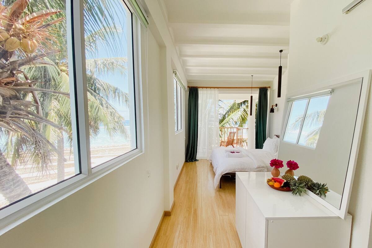 Habitación de hotel con una cama, una mesita con frutas y flores, vistas a palmeras y un balcón con una silla.