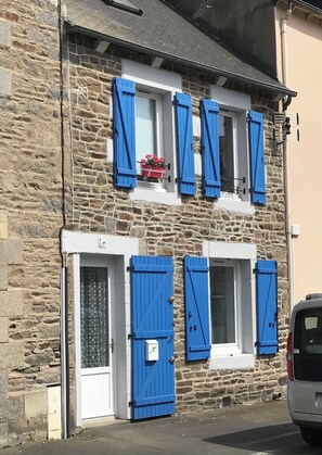Un edificio de piedra con contraventanas azules, una puerta blanca y un pequeño escaparate con flores.