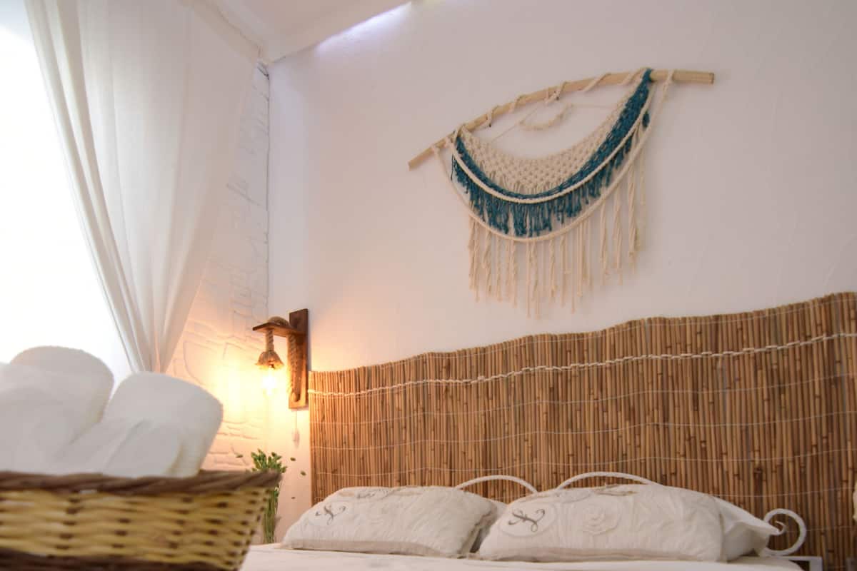 A neatly made bed with a basket of towels, a wall-mounted macrame piece, and a bamboo headboard.