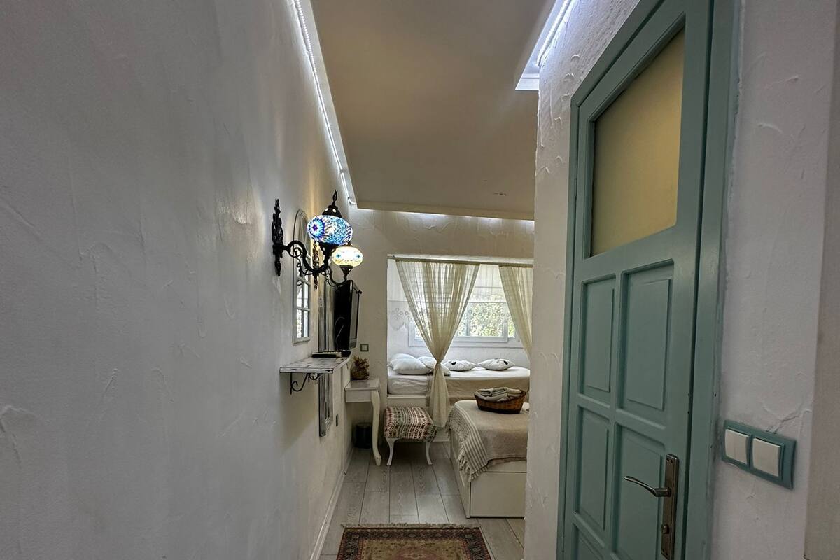 A narrow hallway with a wooden floor leading to a room with a bed and a window with sheer curtains.