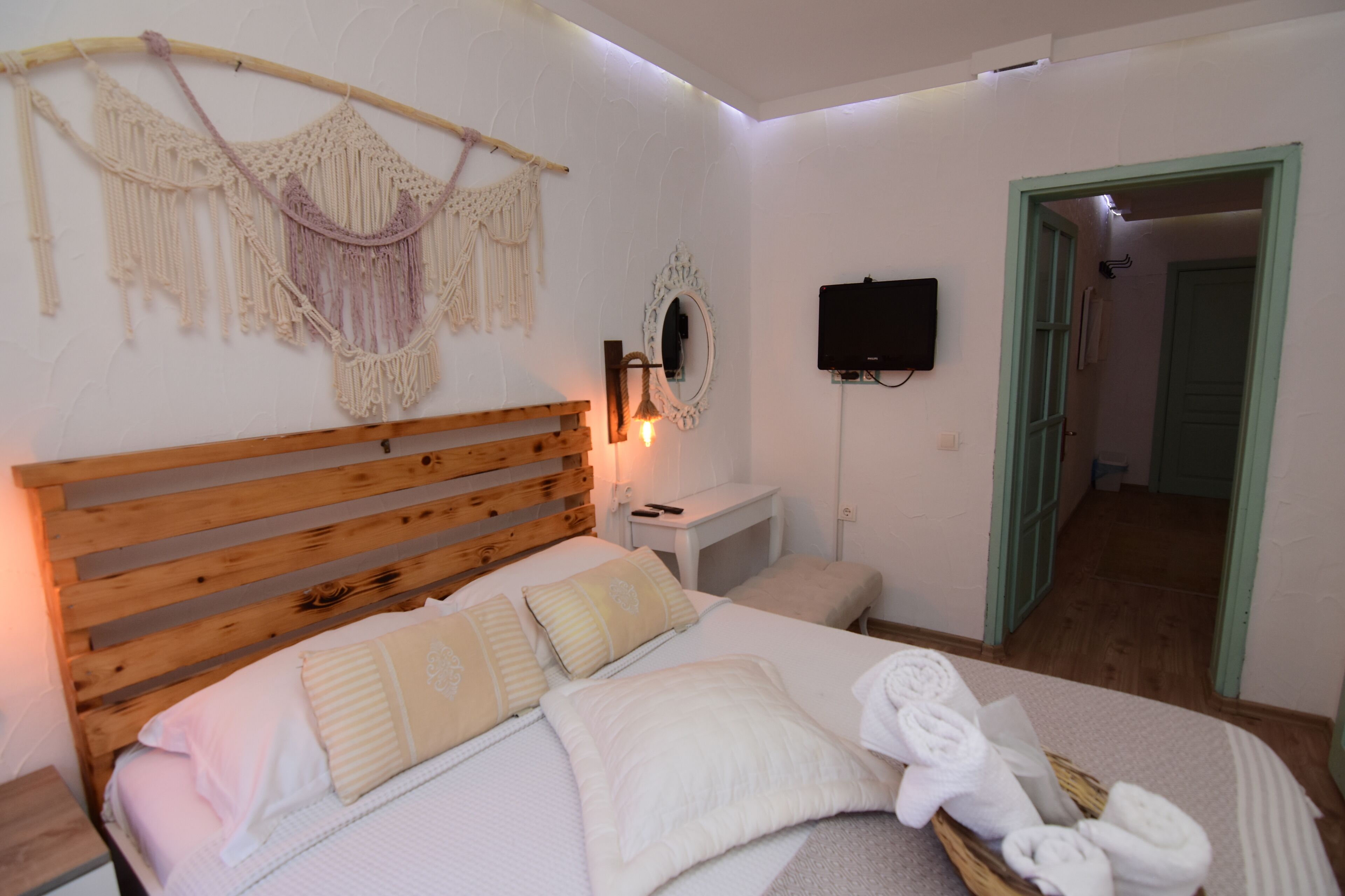 Een slaapkamer met een houten hoofdbord, een bed met wit beddengoed, een macramé wanddecoratie, een nachtkastje met een lamp en een televisie aan de muur.