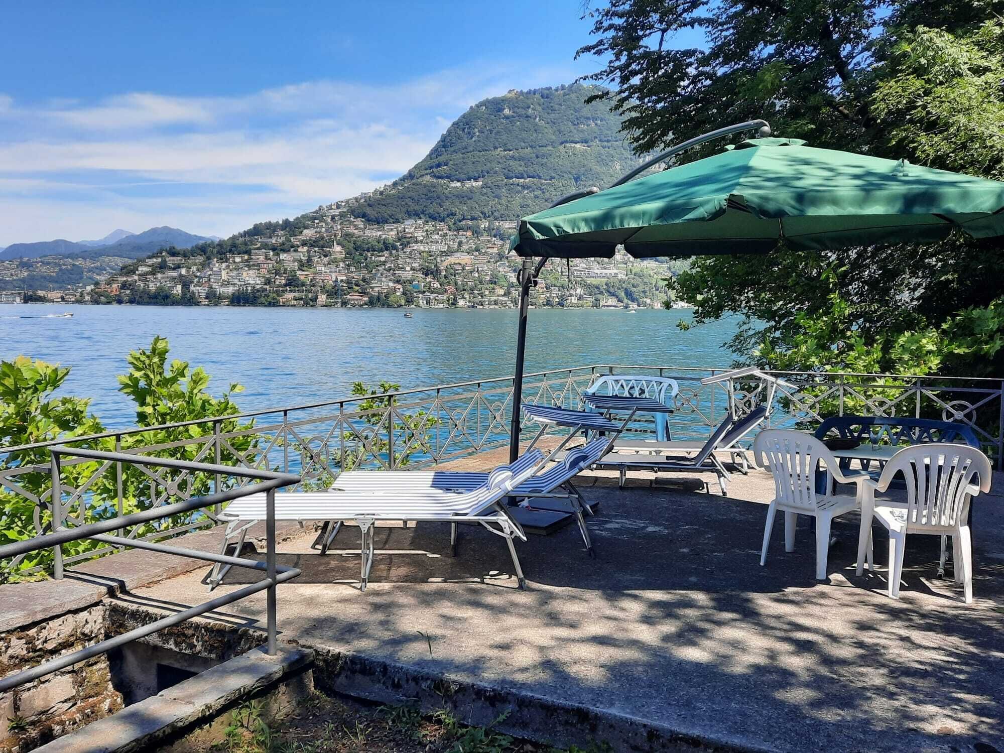 Eine Seeterrasse mit Liegestühlen und einem Tisch unter einem grünen Sonnenschirm, mit Blick auf einen See und eine Hügelstadt.