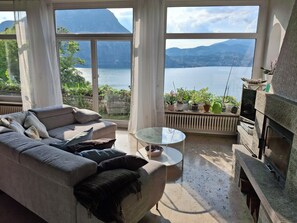 A living room with a sofa, a glass coffee table, and a fireplace. There are potted plants on a windowsill and a view of a lake and mountains through large windows. - Vacation home with an exclusive view of the lake, on Lake Lugano—lakefront property, hiking area. (Lugano)
