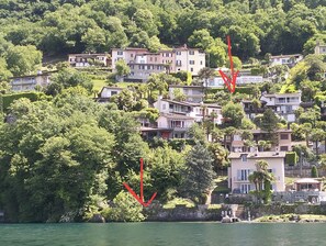 A hillside residential area with houses, some with balconies, and a body of water in the foreground. - Vacation home with an exclusive view of the lake, on Lake Lugano—lakefront property, hiking area. (Lugano)