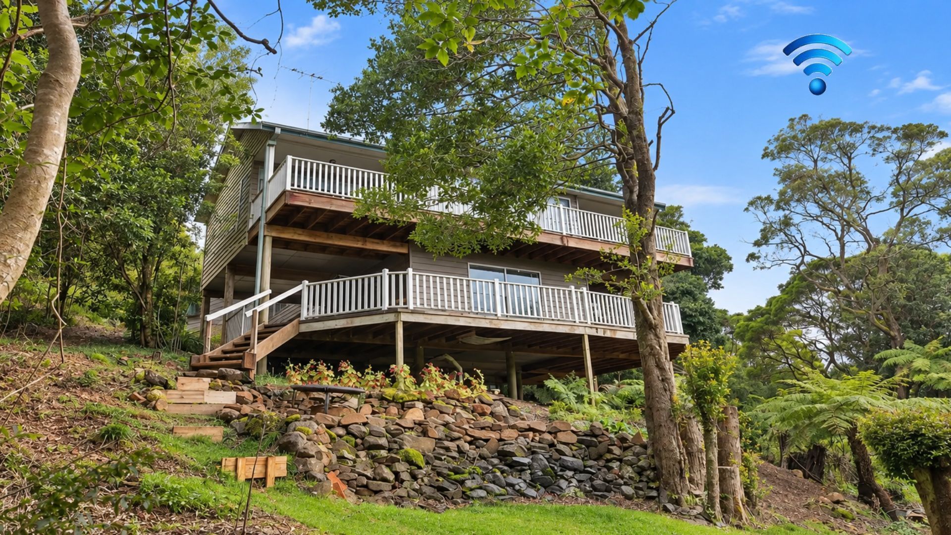 A two-story wooden house with a balcony on each floor, surrounded by trees and a stone wall.