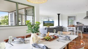 Un comedor moderno con una larga mesa dispuesta para una comida, un sofá al fondo y una cocina visible a través de una puerta abierta.