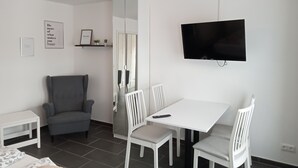 Een moderne, goed verlichte kamer met een eettafel, witte stoelen, een grijze fauteuil, een aan de muur bevestigde televisie en een klein rek met decoratieve objecten.