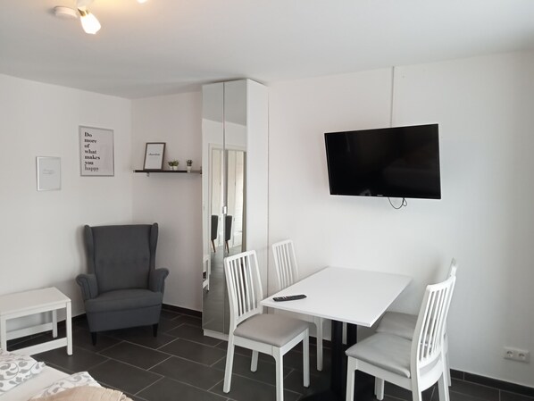 A modern, well-lit room with a dining table, white chairs, a grey armchair, a TV mounted on the wall, and a small shelf with decorative items. - Ferienhaus am Binnenhafen (Emden)