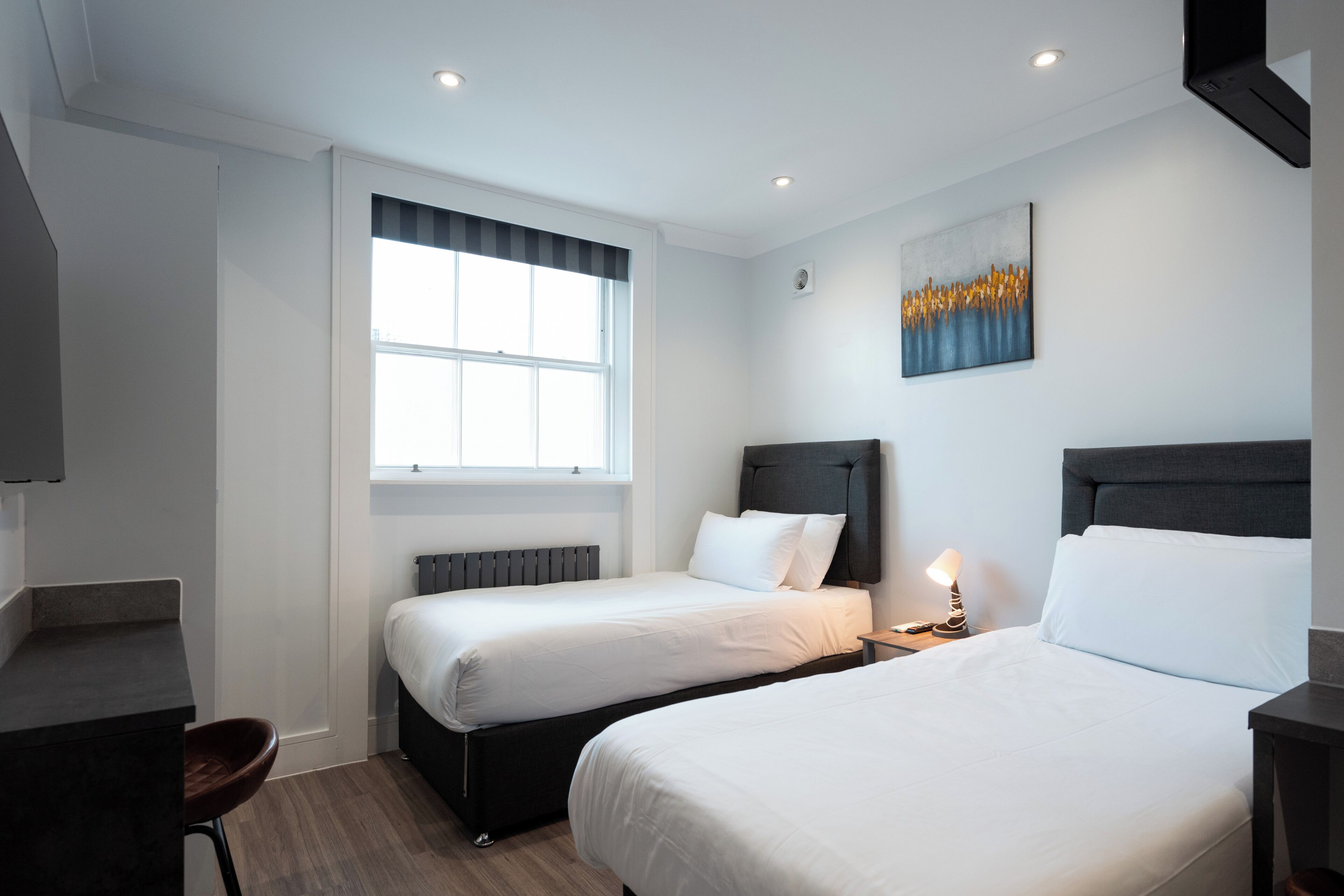 A hotel room with two beds, a desk, a chair, a window, and a painting on the wall.