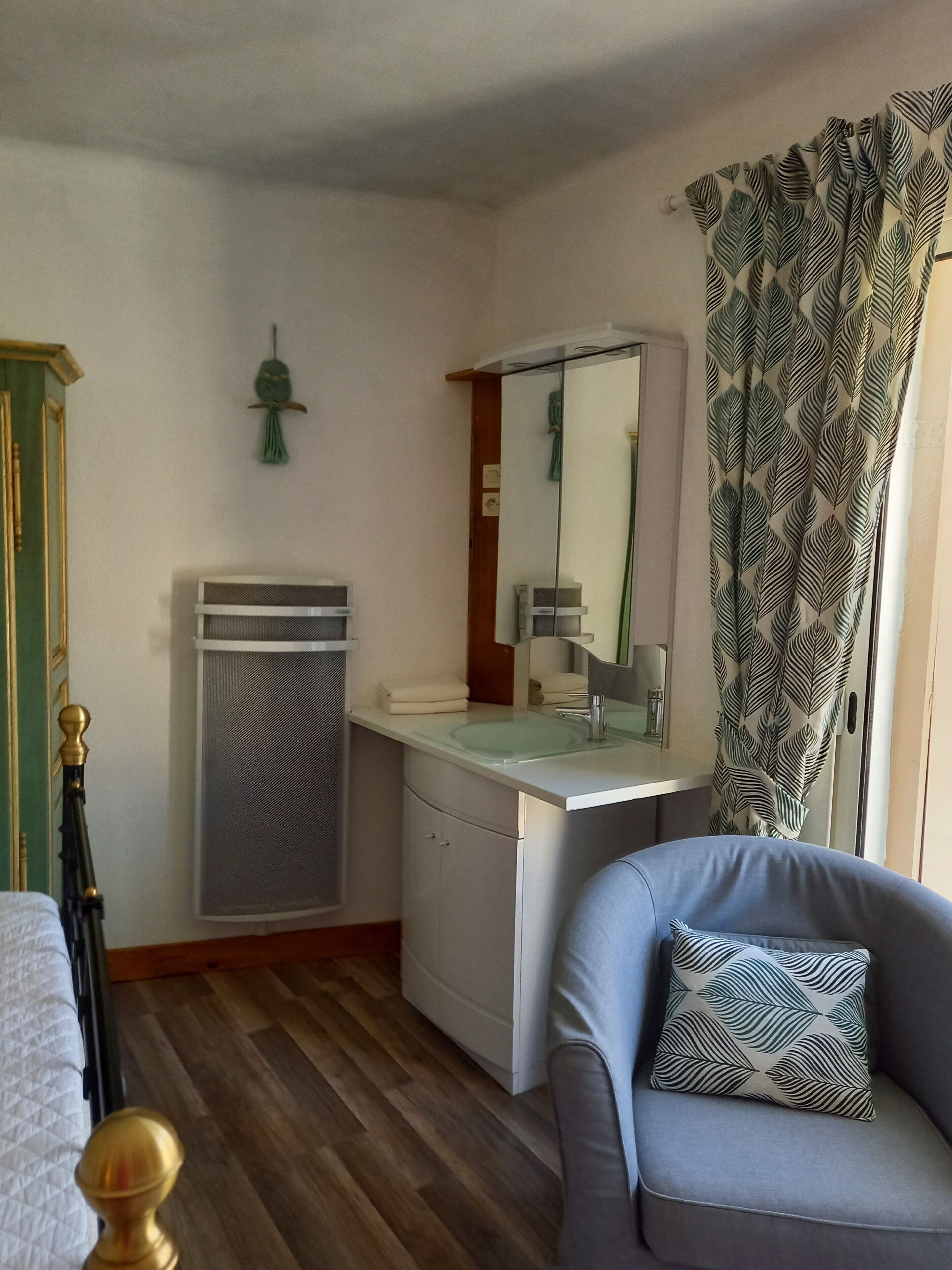 A compact bathroom with a sink, mirror, and toilet. There is a chair with a patterned cushion and a bed with a wooden headboard.