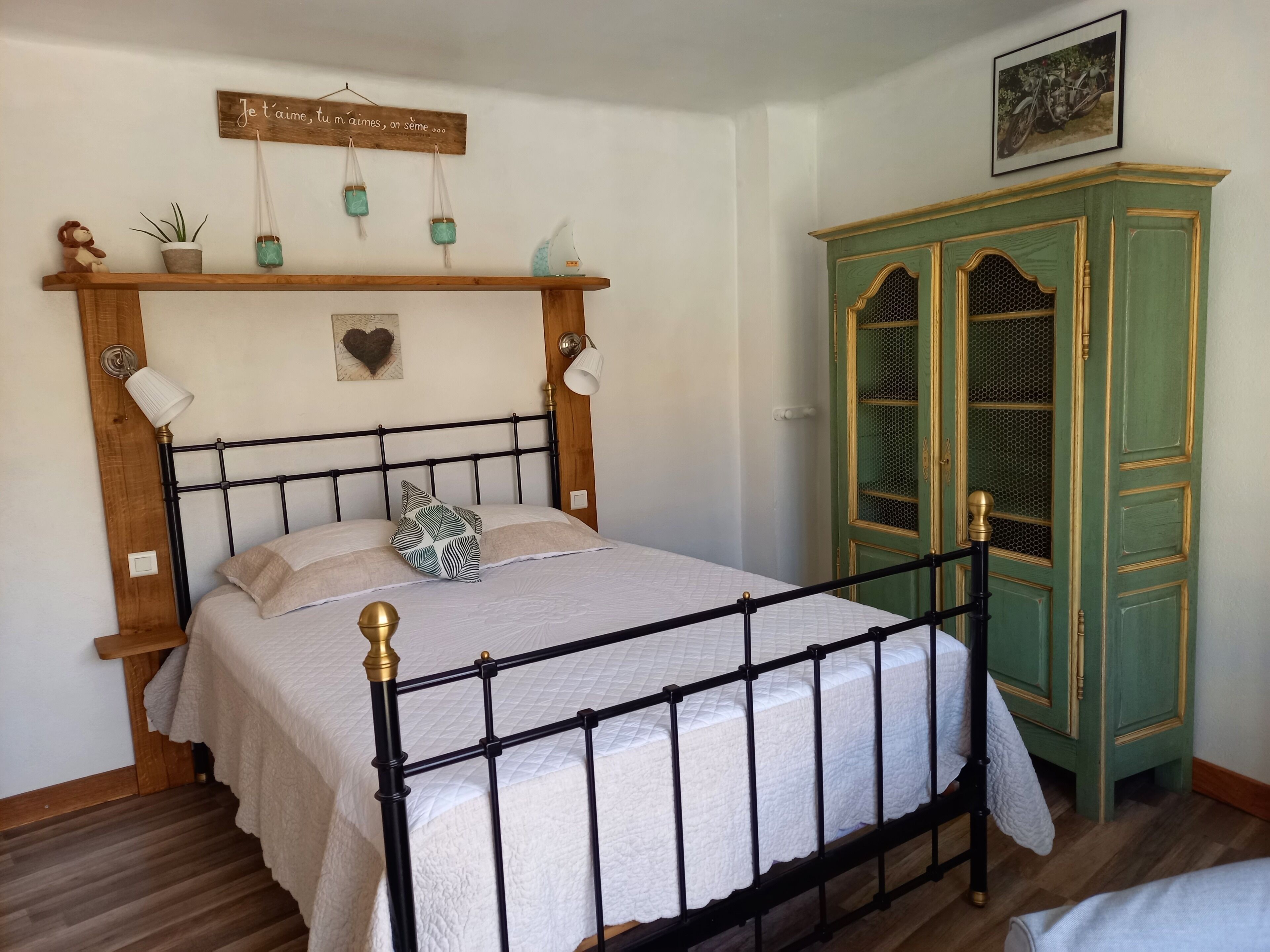 A bedroom with a bed, a wooden headboard, a green cabinet, and a wall-mounted shelf with decorative items.