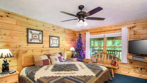 A cozy cabin bedroom with a wooden ceiling fan, a bed with a quilt, a Christmas tree, and a TV on a wooden stand.