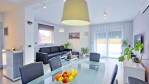 A modern living room with a glass dining table, a sofa, and a large window with blinds.