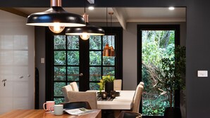 A dining area with a wooden table, two chairs, a coffee table with a vase and a plant, and a window with a view of greenery.