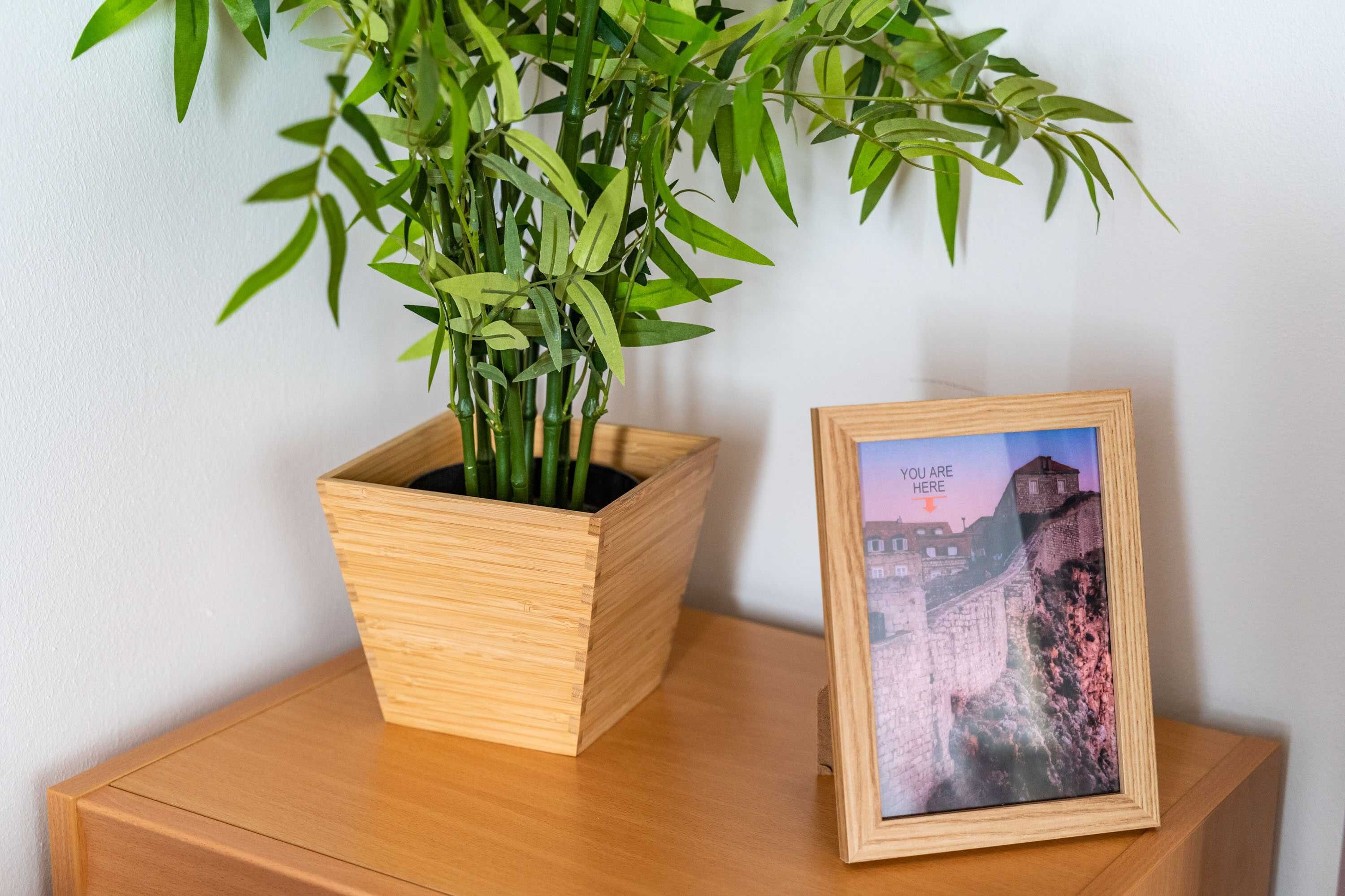 Een houten nachtkastje met een bamboeplant in een vierkante houten plantenbak en een ingelijst beeld van een historisch gebouw.