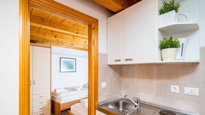 A compact kitchen with a sink, stove, and cabinets. A bed with a white blanket is visible in the adjacent room. - Apartment Ribambit (Dubrovnik)