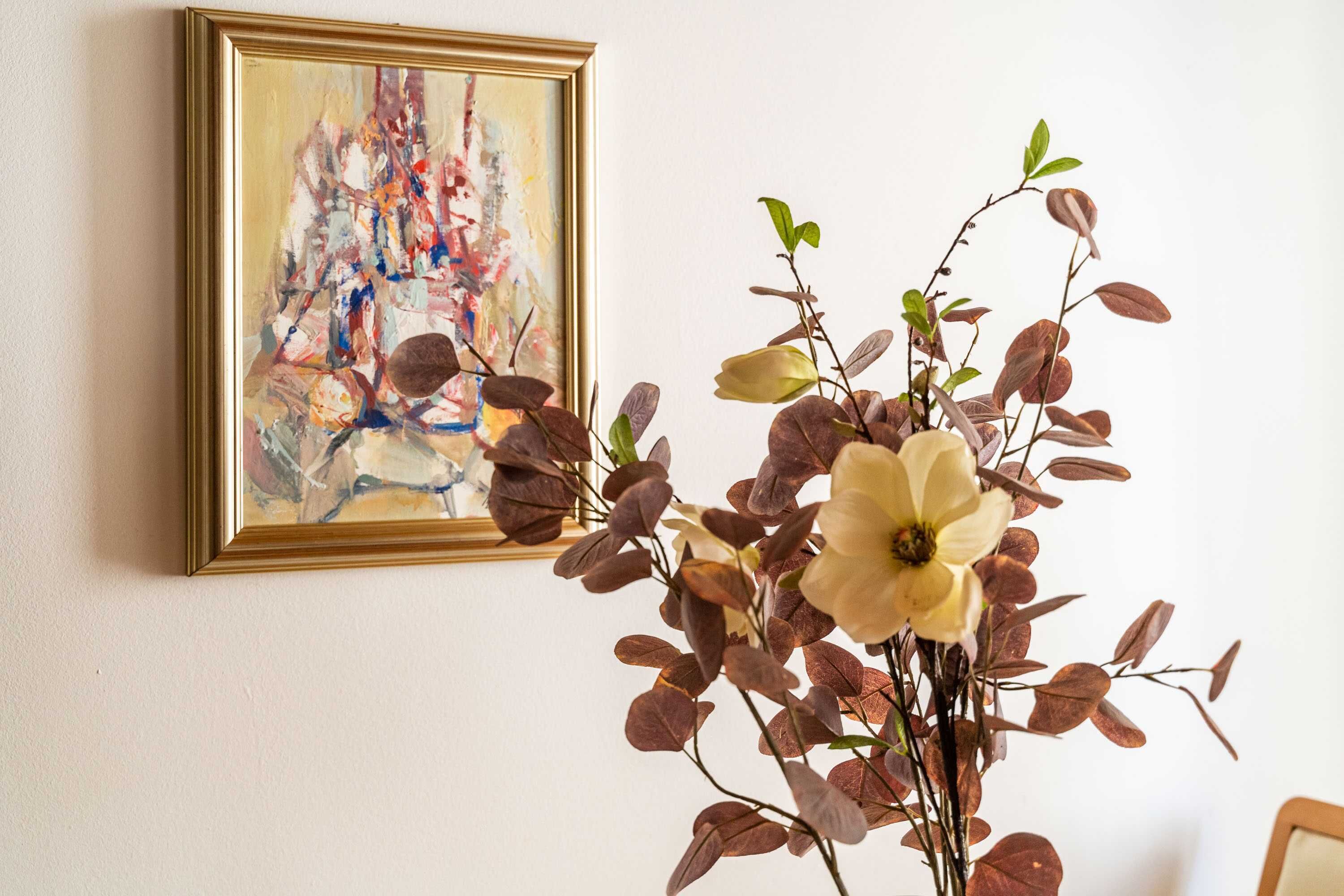A framed abstract painting on a wall, a vase with a white flower, and a plant with brown leaves.