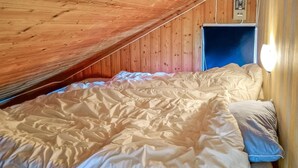 Ein Schlafzimmer mit schrägverdachter Decke, Holzwänden, einem Bett mit weißer und beiger Bettdecke sowie einem Fenster mit Vorhängen.