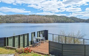 Une terrasse en bois avec une table et des chaises, donnant sur un lac paisible, le tout entouré de collines boisées.