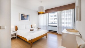 Une chambre d’hôtel avec un lit en bois, un bureau, une chaise, une lampe, un tableau au mur et une fenêtre avec des rideaux.