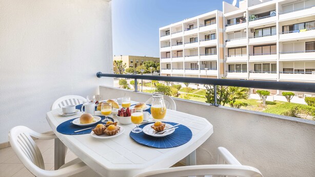 Un balcon avec une table préparée pour le petit-déjeuner, comprenant des assiettes de nourriture, un pichet de jus d’orange et un saladier de fruits.
