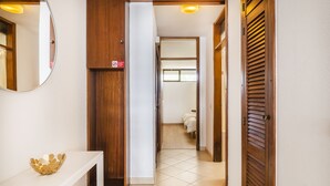 Un couloir avec une console blanche, un miroir et une porte en bois donnant sur une pièce où se trouve un lit.