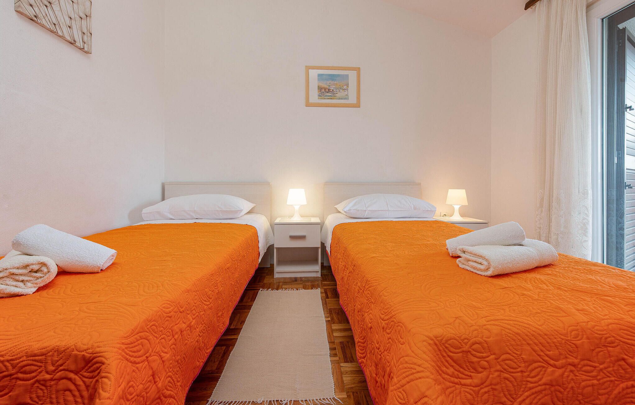 Hotellihuoneessa on kaksi sänkyä, joissa on oranssit päiväpeitteet, valkoiset tyynyt ja rullalle taitetut pyyhkeet. Huoneessa on myös kaksi yöpöytää, joissa on lamput, seinällä on kehystetty kuva ja ikkunassa on verhot.