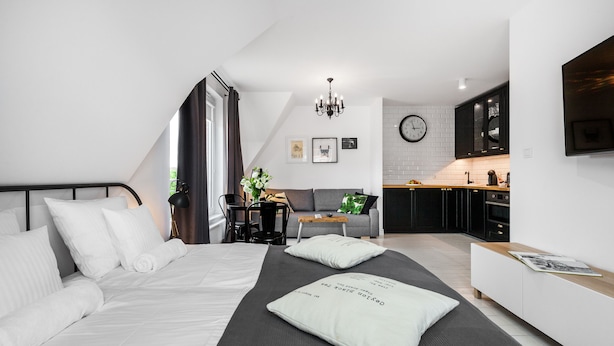 A modern bedroom with a large bed, a small desk, and a kitchenette in the background.