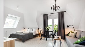 A modern bedroom with a bed, a sofa, a table, and a chair. There is a window with curtains, a chandelier, and a view of greenery outside.