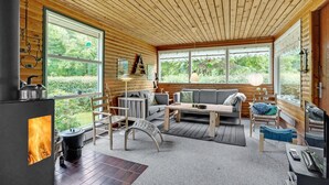 A cozy cabin interior with a fireplace, wooden walls, a sofa, and large windows offering a view of the greenery outside. - Amazing home in Nykøbing M (Nykøbing M)