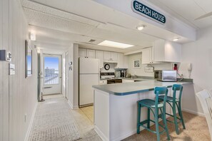 Une cuisine dotée d’un îlot central, de meubles blancs et offrant une vue sur la plage grâce à une porte ouverte.