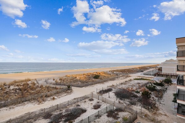 Une vue sur la plage avec une clôture en bois, un sentier sablonneux et un bâtiment doté de balcons.