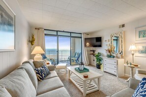 Un salon spacieux doté d’une grande fenêtre donnant sur la plage, d’un téléviseur à écran plat et d’un coin salon confortable avec un canapé et des fauteuils.