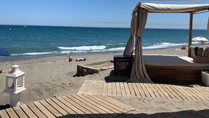Een houten terras aan het strand, met een overdekte zithoek en een witte parasol.