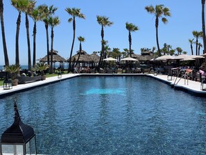 A swimming pool with lounge areas, palm trees, and thatched umbrellas.