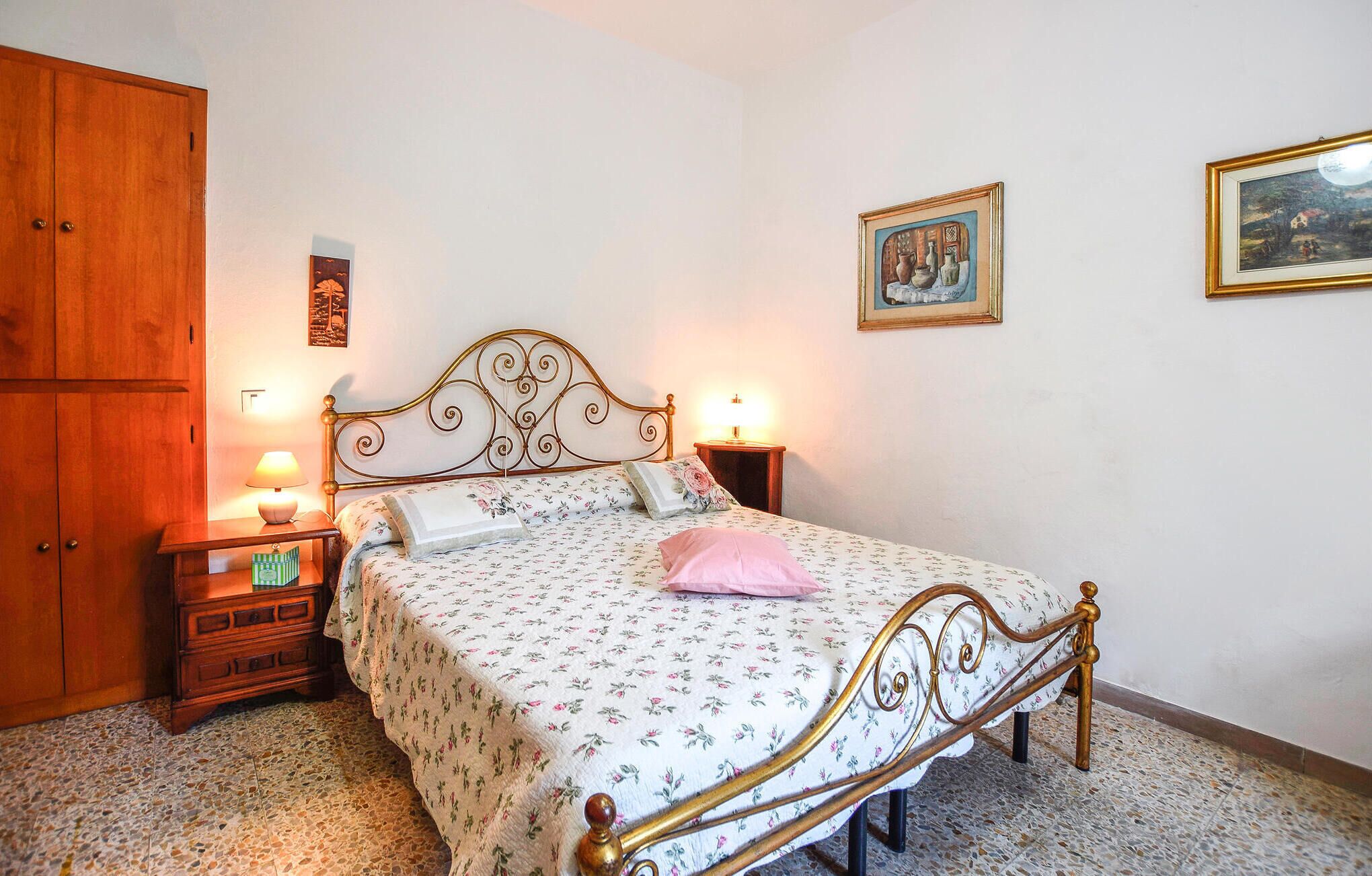 A bedroom with a bed, wooden wardrobe, nightstands, and wall-mounted paintings.